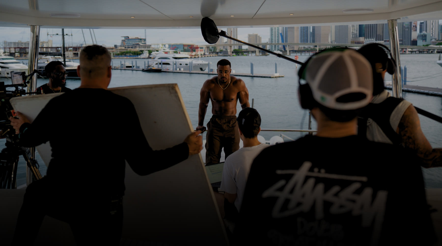 Behind the scenes on the #ALLIN launch shoot: Ashton Hall on a boat deck facing the camera crew and a boom mic, with the Miami skyline behind him.