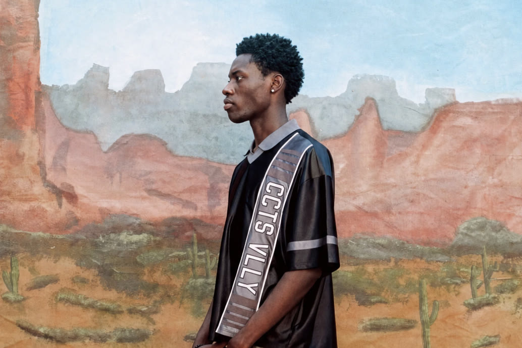 A model in profile wearing an AZVA jersey and grey sweats against a painted desert backdrop.